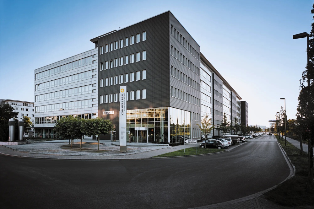 Modern office building with a glass facade, surrounded by parked cars and trees, under a blue sky.