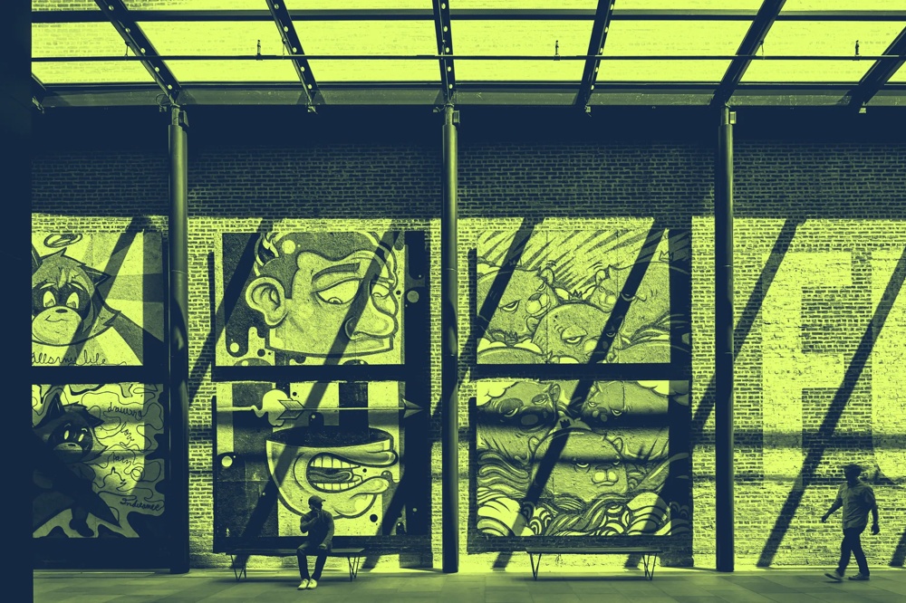 Wandgemälde mit Cartoon-Gesichtern in einer Halle, zwei Personen davor, Schatten von Glasdach fallen auf die Wand.