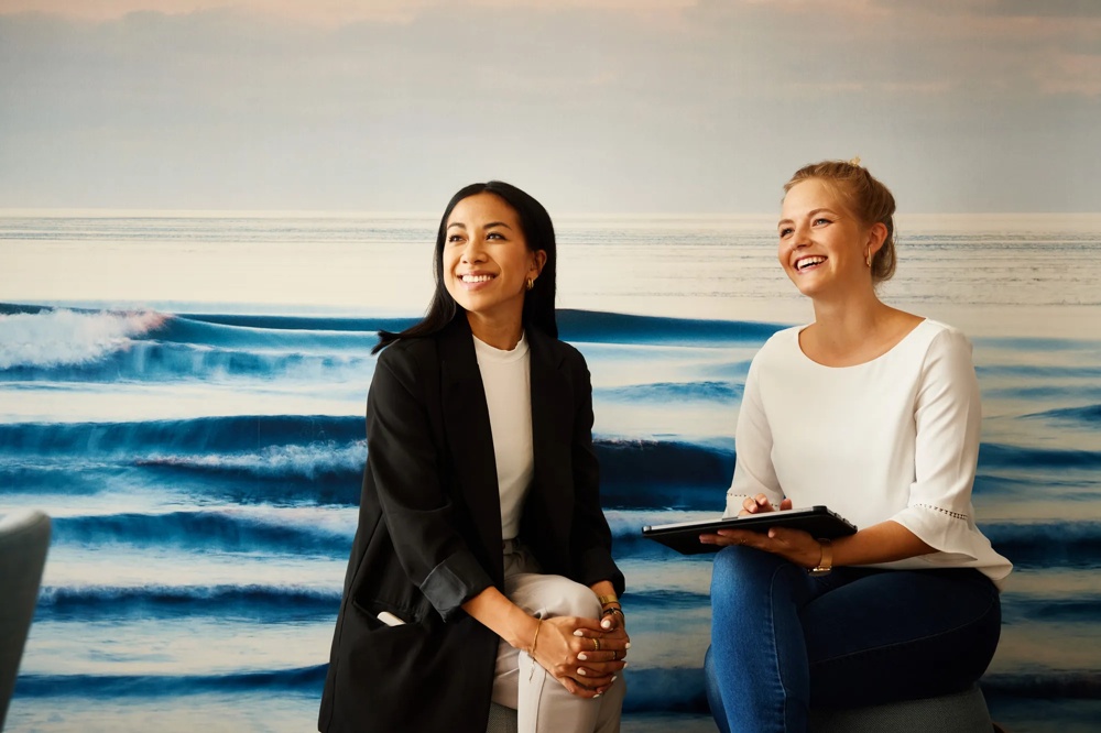 Two smiling women sitting in front of a wall painting with ocean waves, one holding a tablet.