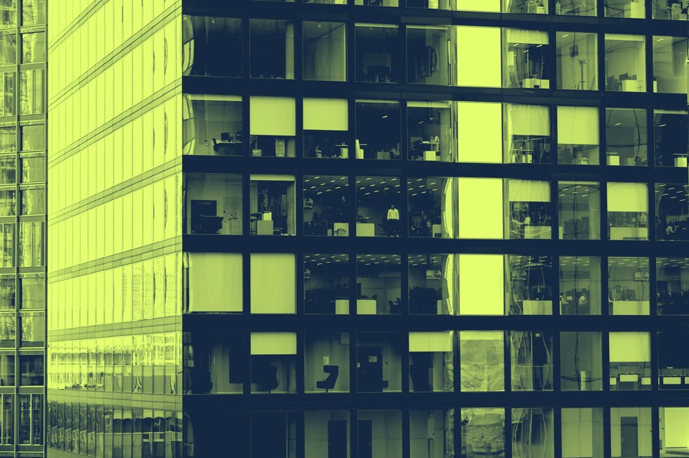 Ein modernes Bürogebäude mit großen, rechteckigen Fenstern, durch die Büros und Menschen sichtbar sind.