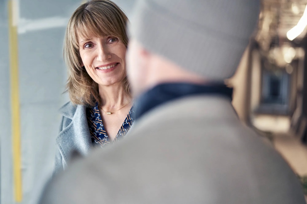 Eine Frau in grauem Mantel lächelt einen Mann mit Mütze an, unscharfer Hintergrund.