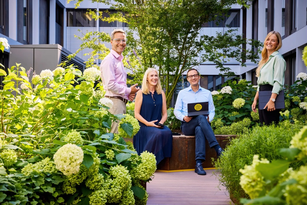 Four people smiling in a green garden in front of a modern building.