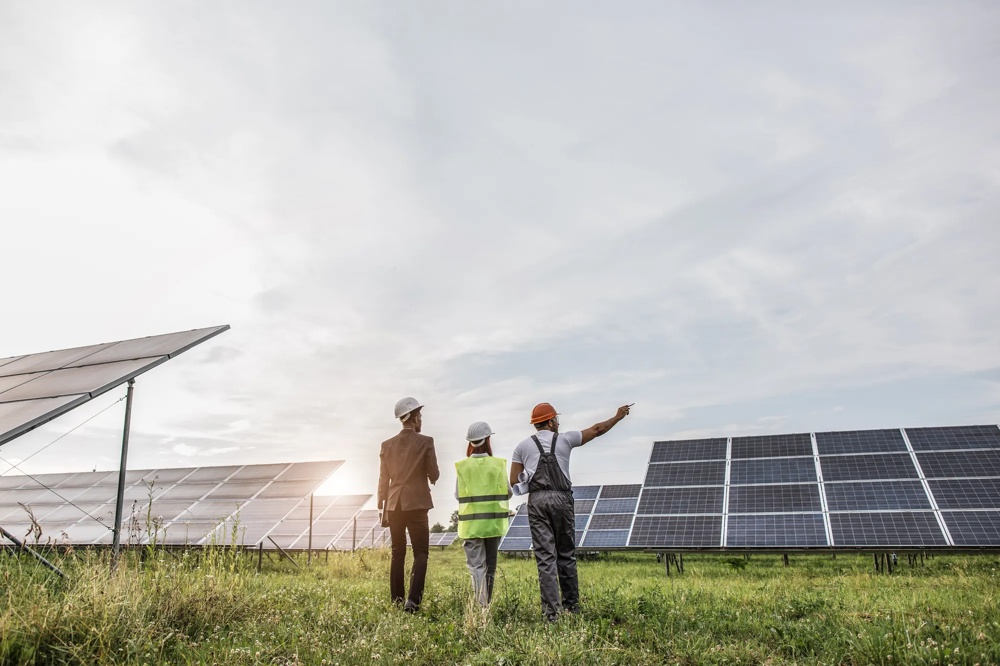 Drei Personen mit Helmen und Westen stehen vor Solarpanels auf einem Feld.