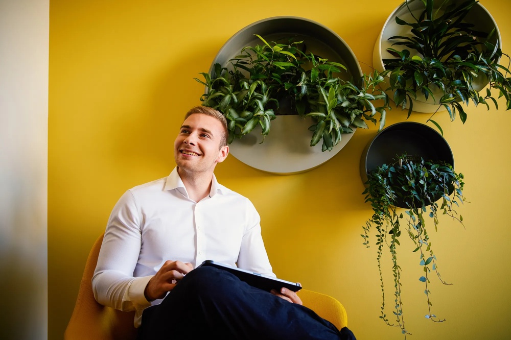 A smiling man in a white shirt sitting in front of a yellow wall with green plants in round pots.