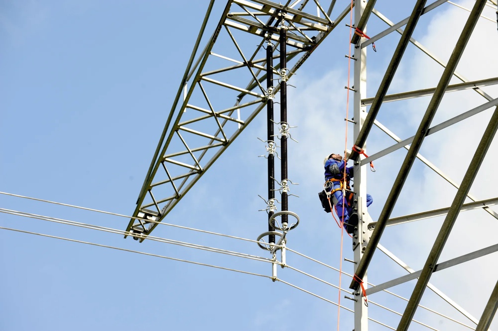 Ein Arbeiter in blauer Schutzkleidung klettert an einem Strommast bei klarem Himmel.
