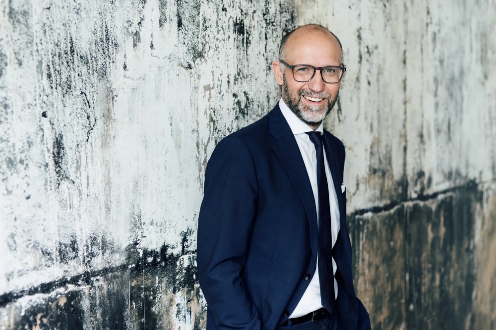 A man in a suit standing smiling in front of a textured white wall.