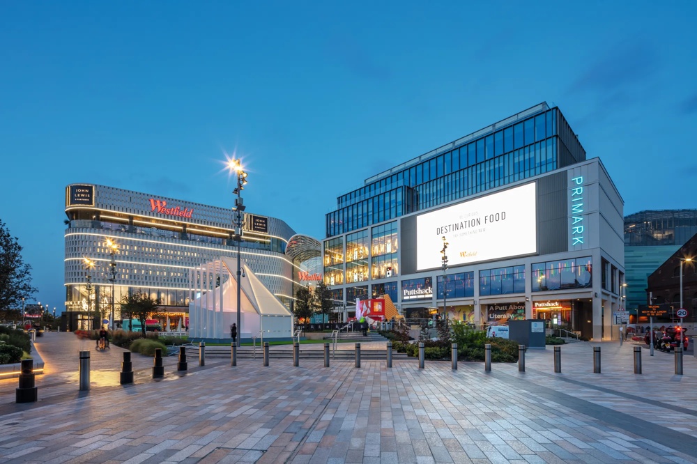 Einkaufszentrum bei Dämmerung, beleuchtete Schilder, moderner Platz mit Bäumen und Laternen, klarer Himmel.