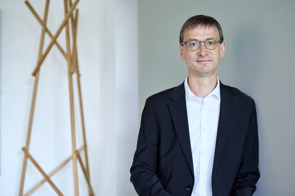 Ein Mann mit Brille im Anzug steht vor einer grauen Wand und schaut freundlich in die Kamera.
