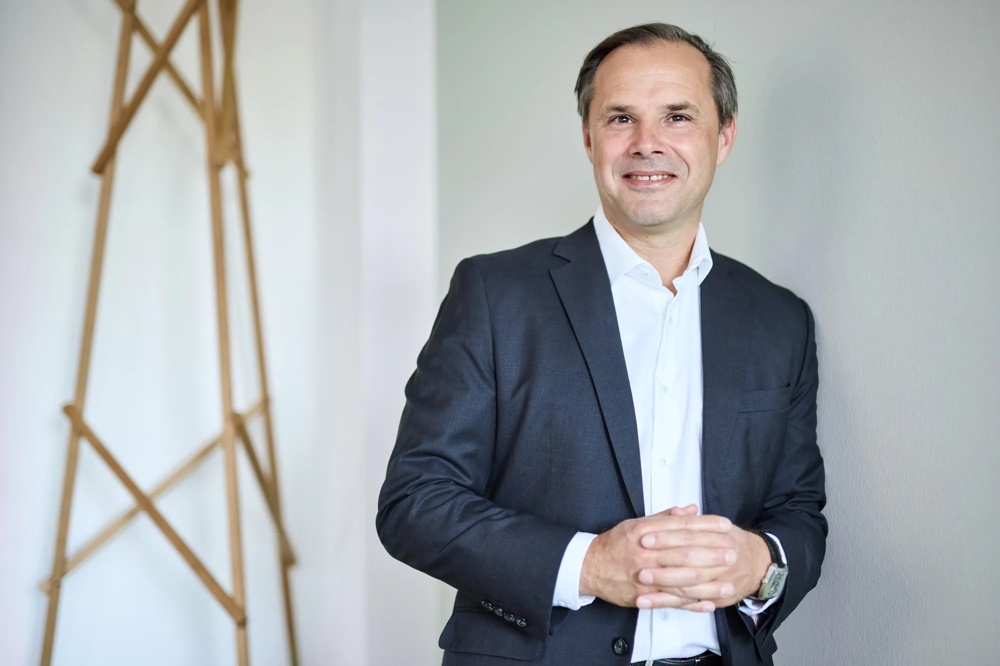 Ein Mann in Anzug lächelt vor einem neutralen Hintergrund.