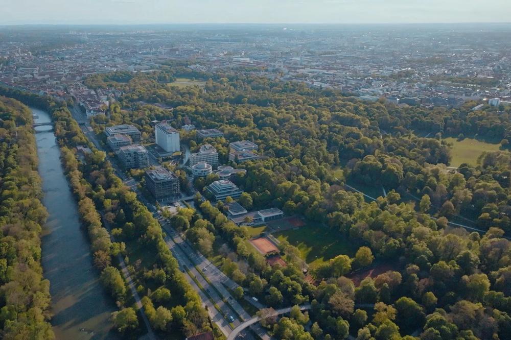 Luftaufnahme einer Stadt mit Fluss, umgeben von dichten, grünen Bäumen und Gebäuden im Hintergrund.