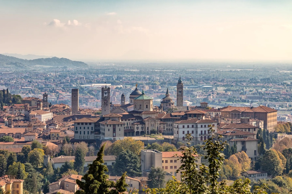 Panorama der Altstadt von Bergamo mit roten Ziegeldächern, historischen Türmen und sanften Hügeln im Hintergrund.