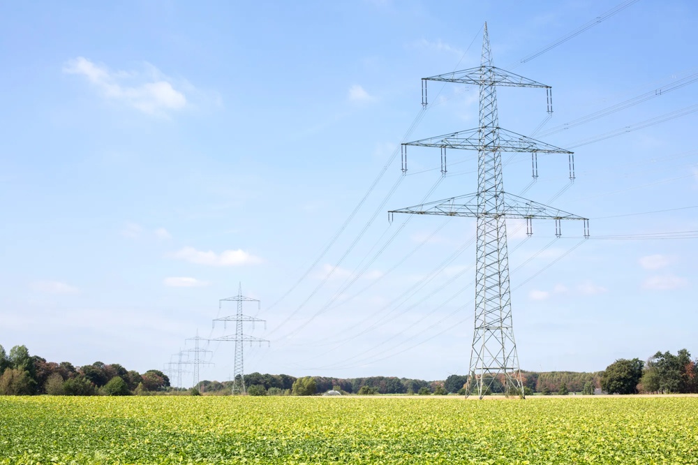 Strommasten stehen auf einem weiten Feld unter blauem Himmel mit vereinzelten Wolken.