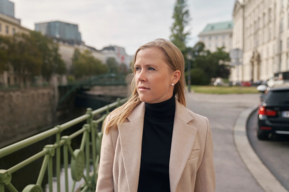 Frau in beigem Mantel steht auf einer Brücke, Stadtgebäude und Autos im Hintergrund.