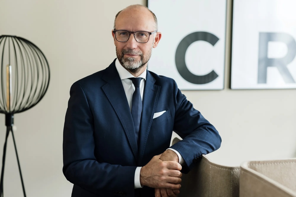 Ein Mann im Anzug mit Brille lehnt an einem Stuhl, Hintergrund mit einem großen Buchstaben C.