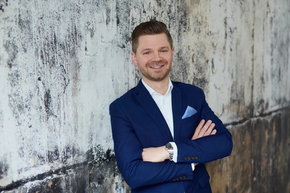 Ein Mann im blauen Anzug steht lächelnd mit verschränkten Armen vor einer rauen, gemusterten Wand.