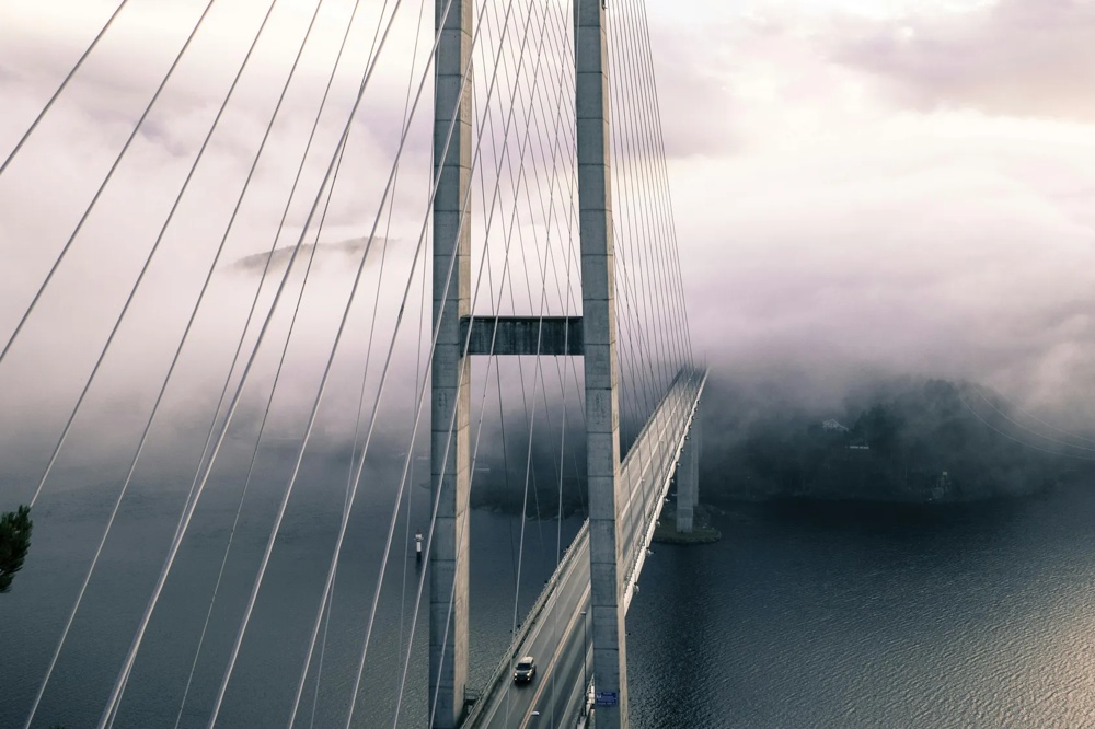 Eine lange Hängebrücke führt über nebliges Wasser, umgeben von dichtem Nebel.