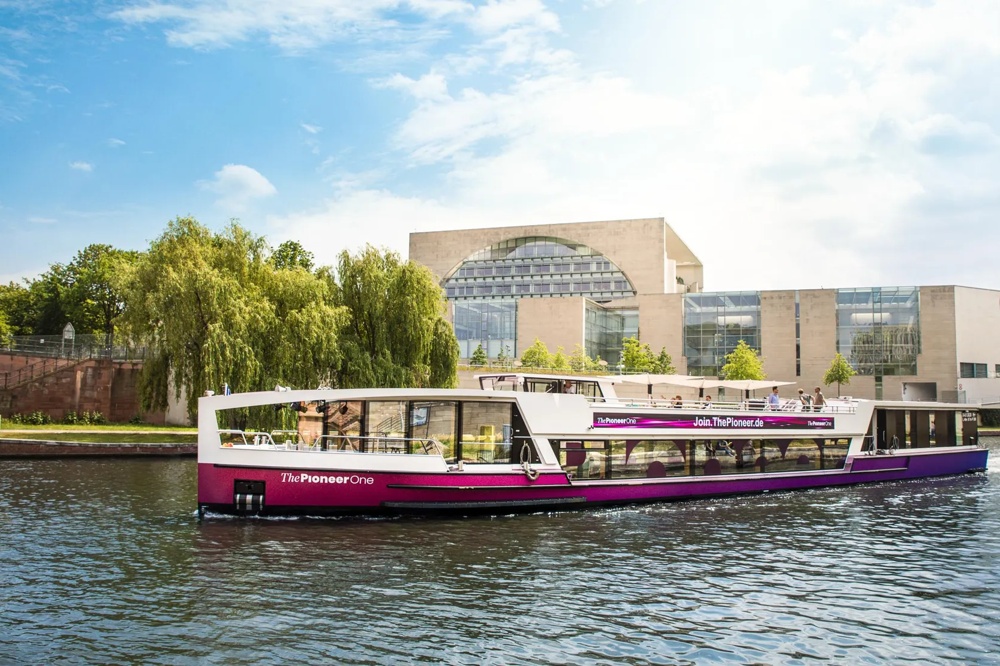 Ein modernes Ausflugsboot fährt auf einem Fluss vor einem großen, modernen Gebäude und grünen Bäumen.