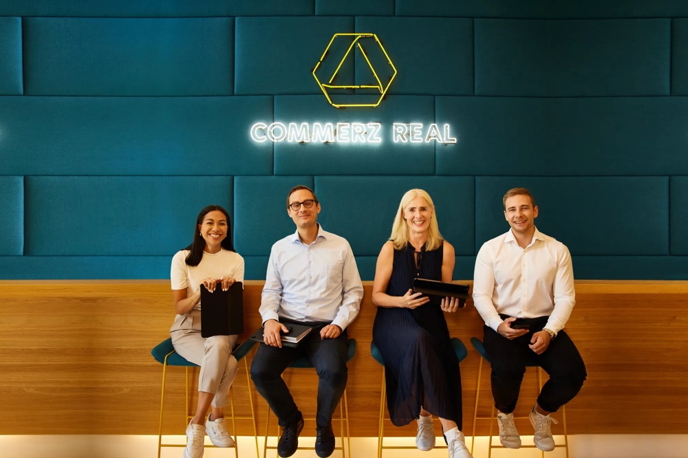 Four people are sitting on bar stools with a smile in front of a blue wall with the “Commerz Real” logo.