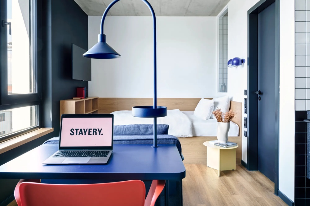 Ein modernes Zimmer mit Laptop auf blauem Tisch, Bett, Vase, und blauer Lampe.