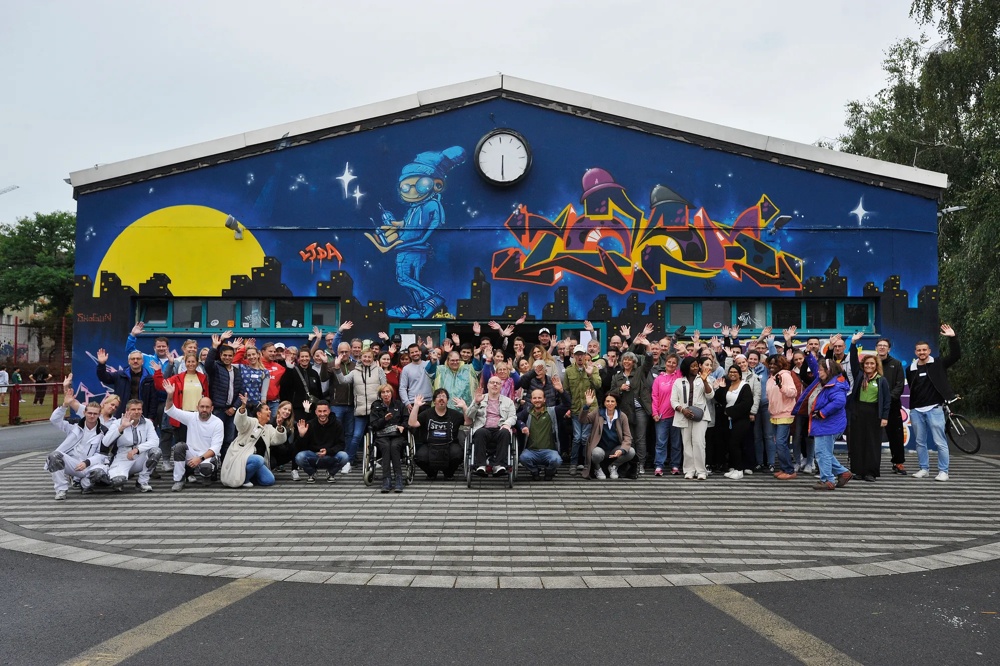 A large group of people posing in front of a building with colourful graffiti and a clock.