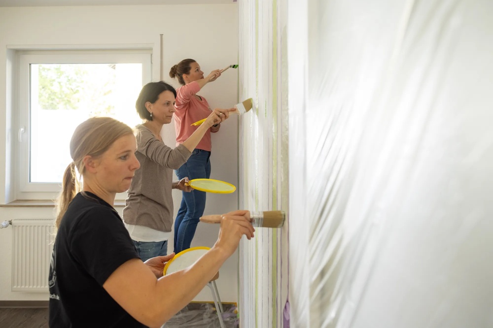 Three women painting a wall with brushes and yellow paint bowls.