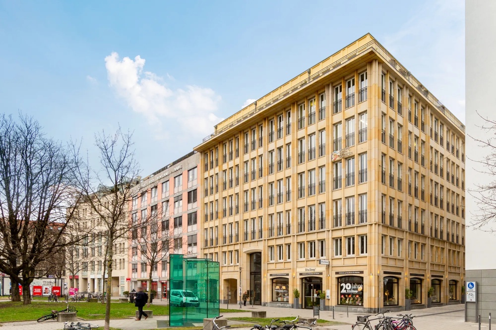 Ein mehrstöckiges, beiges Gebäude mit vielen Fenstern, umgeben von Bäumen und Fahrrädern, unter blauem Himmel.
