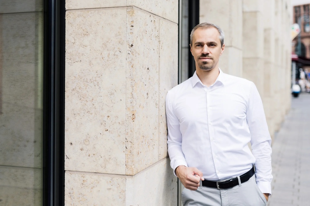 Ein Mann in weißem Hemd lehnt an einer beigefarbenen Steinwand in einer städtischen Umgebung.