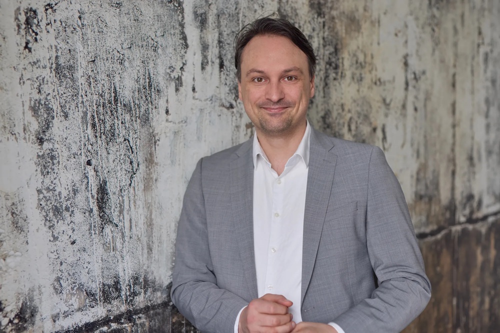 Ein Mann im grauen Anzug steht vor einer strukturierten, verwitterten Wand und lächelt in die Kamera.