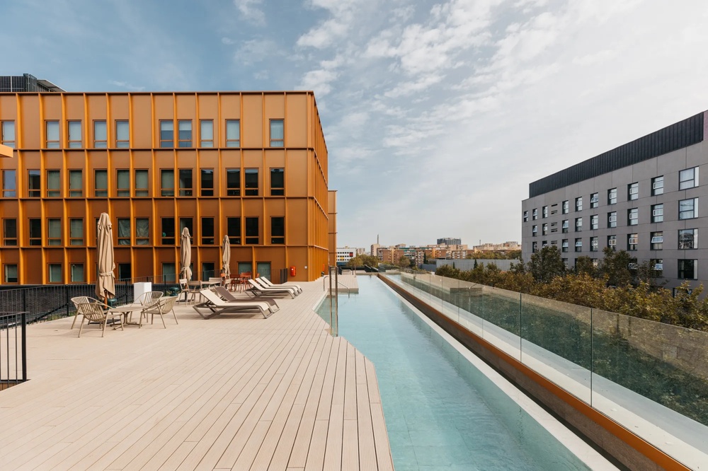Dachterrasse mit Liegestühlen, Sonnenschirmen und Pool neben einem modernen, orangefarbenen Gebäude.