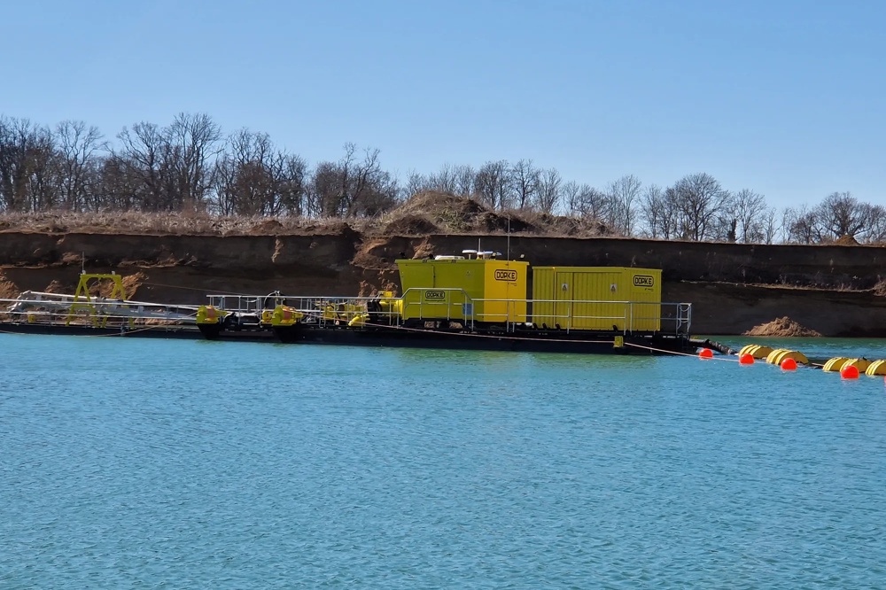 Gelbes Industriegebäude auf einem Floß im blauen Wasser, mit kahlen Bäumen im Hintergrund.