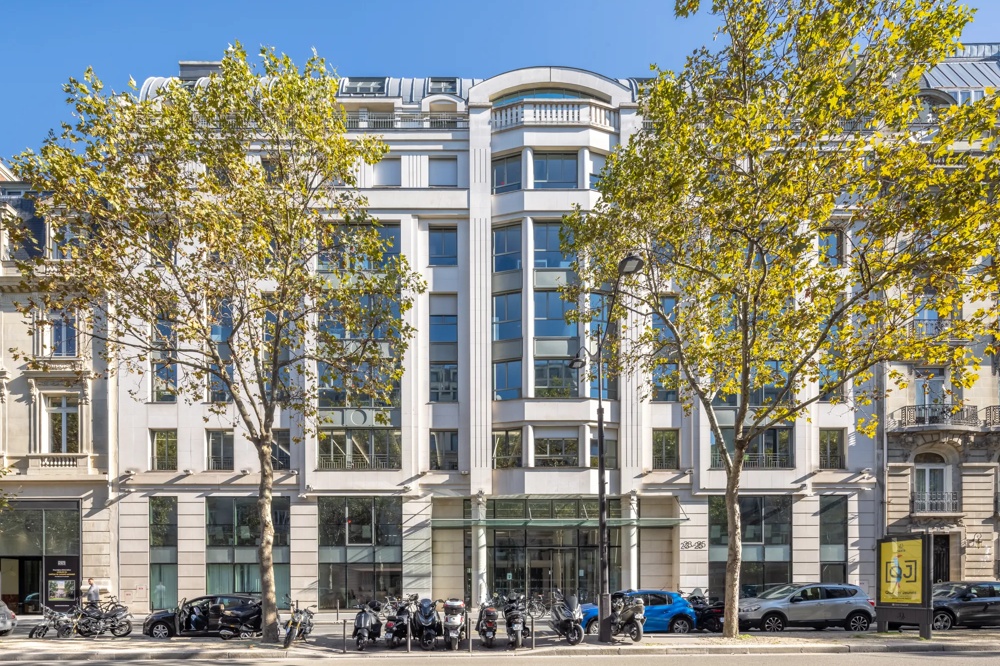 White facade of a modern building surrounded by green trees under a blue sky.