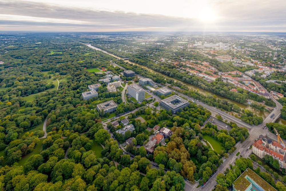 Luftaufnahme eines weitläufigen Parks mit vielen Bäumen, Gebäuden und einem Fluss unter bewölktem Himmel.