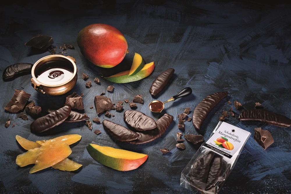 Schokolierte Mangostreifen und frische Mango auf dunklem Untergrund, umgeben von Schokoladenstücken.