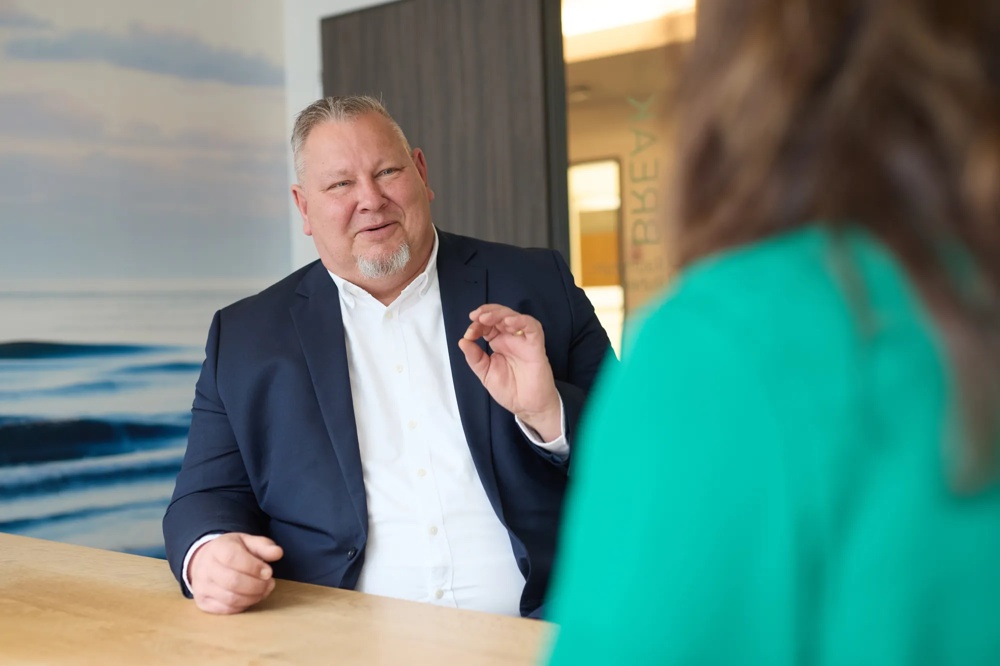 Es wird der Commerz Real Mitarbeiter Claus André Krüger, Senior Key Account Manager für Sales Private and Business Clients SW, dargestellt. Er sitzt am Tisch, spricht mit einer Person im Vordergrund und formt mit den Fingern ein erklärendes Handzeichen.