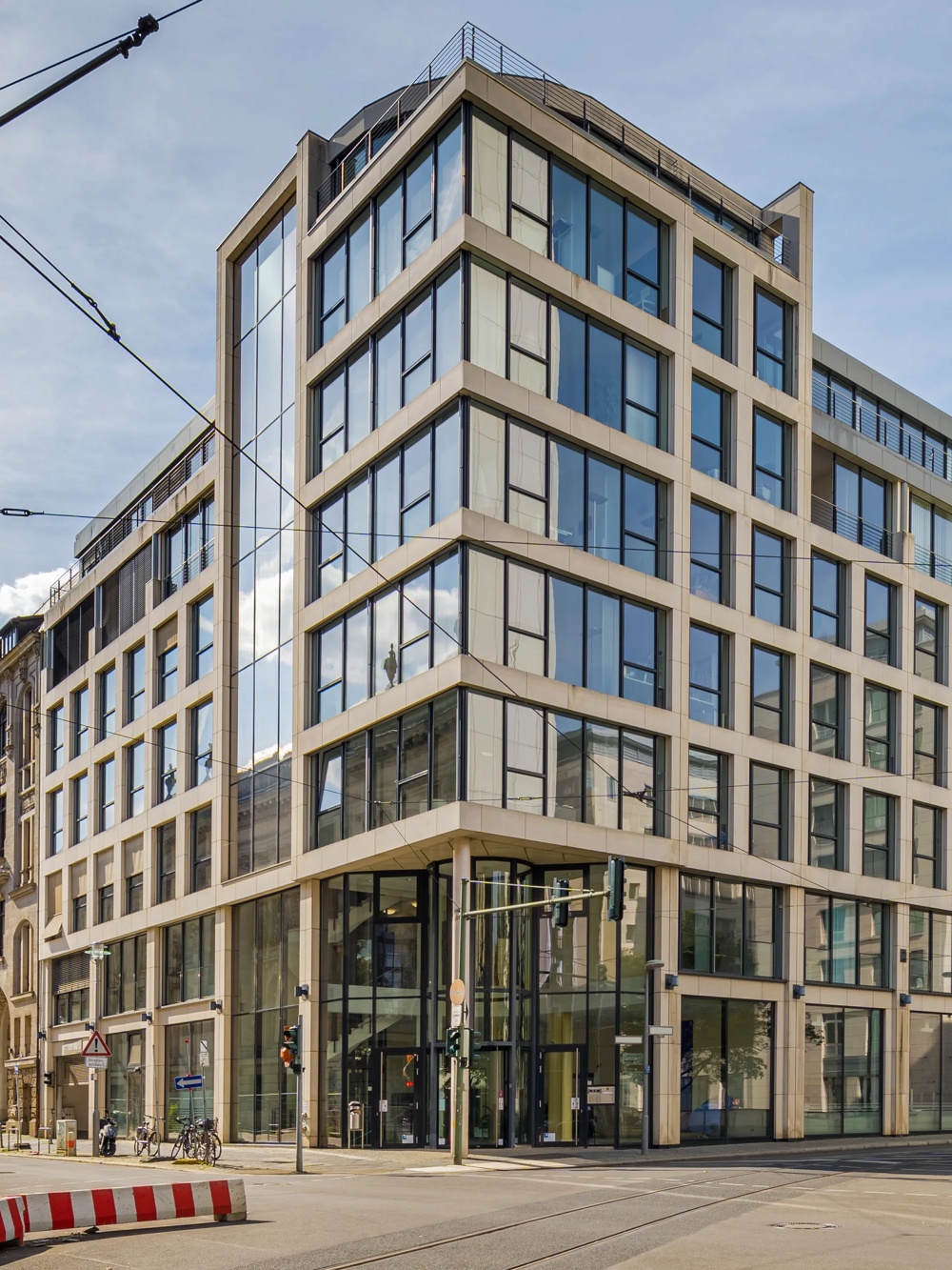 Modernes Eckgebäude mit Glasfassade an einer Straßenecke, umgeben von historischen Gebäuden bei klarem Himmel.