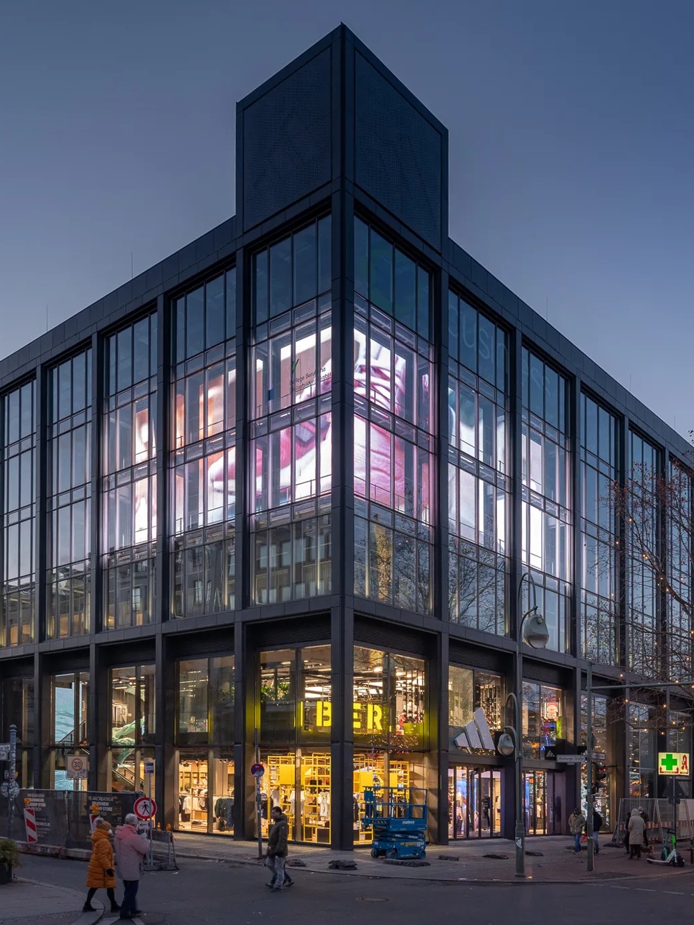 Modernes Glasgebäude bei Dämmerung, leuchtende Werbeanzeigen, Menschen und Bus auf belebter Straße.