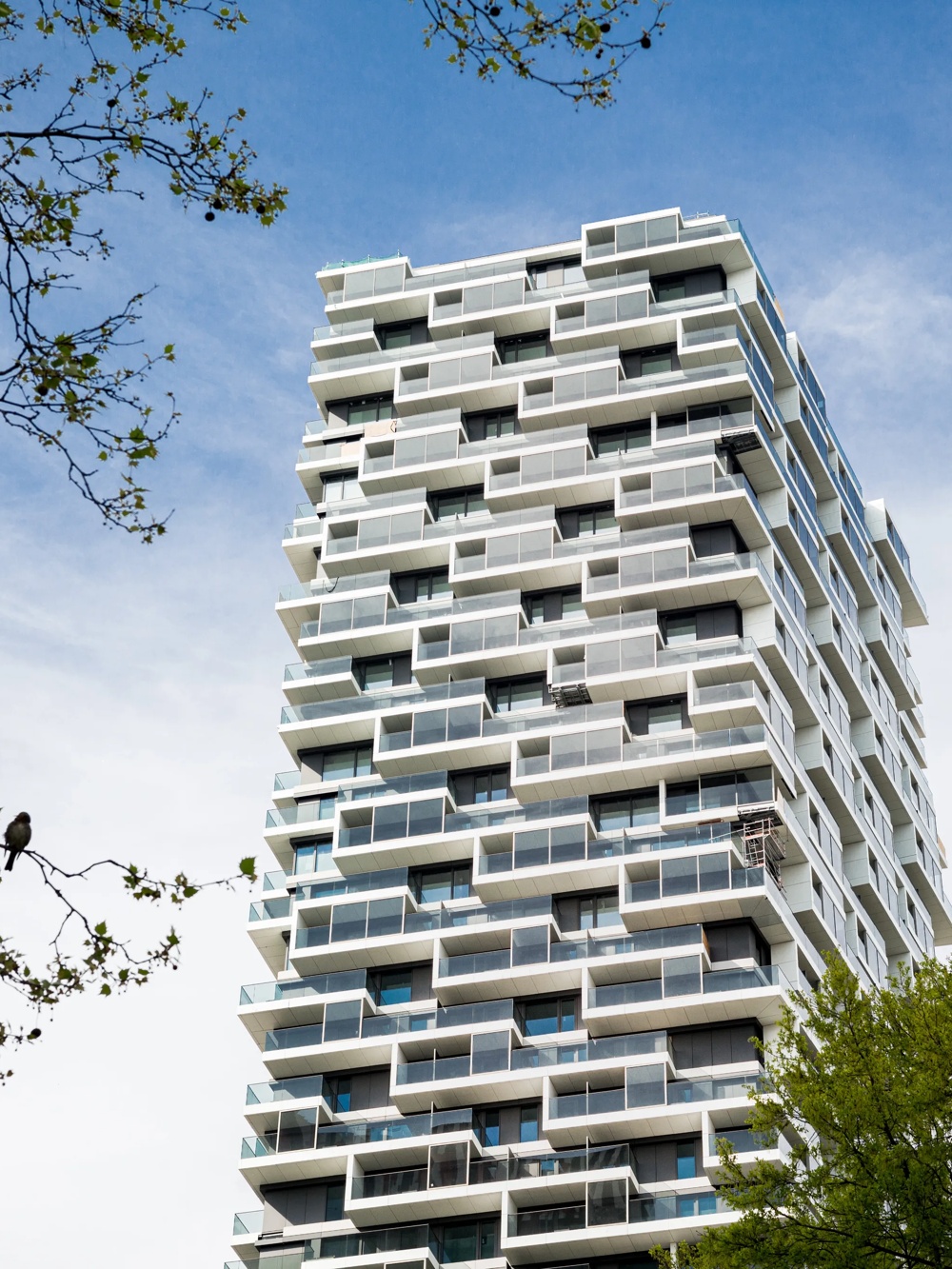 Ein modernes Hochhaus mit versetzten Balkonen, umgeben von grünen Bäumen und blauem Himmel.