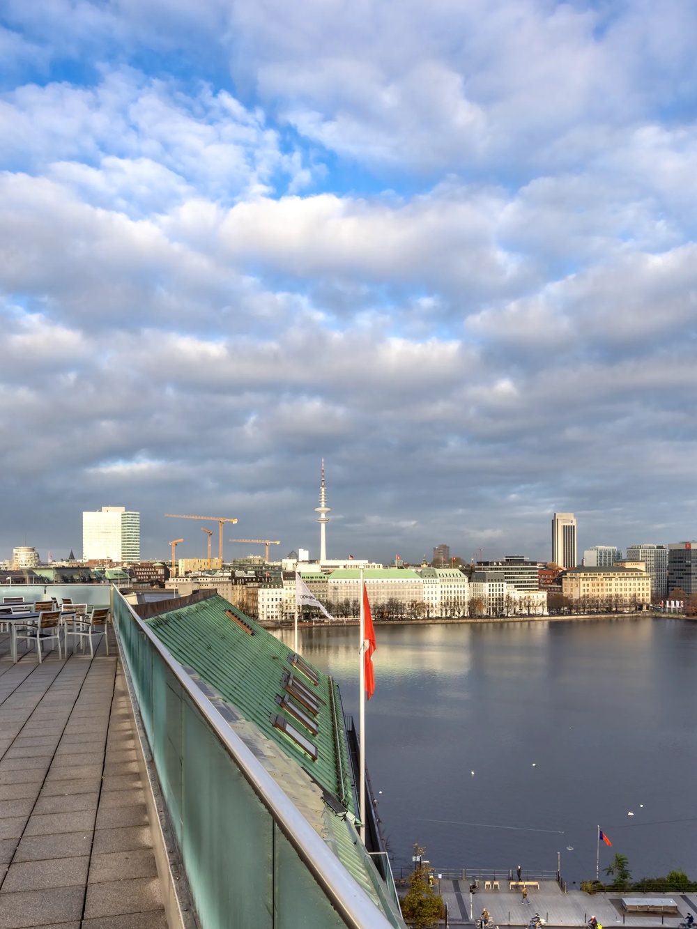 Blick von einer Dachterrasse auf einen See, umgeben von Gebäuden unter bewölktem Himmel.