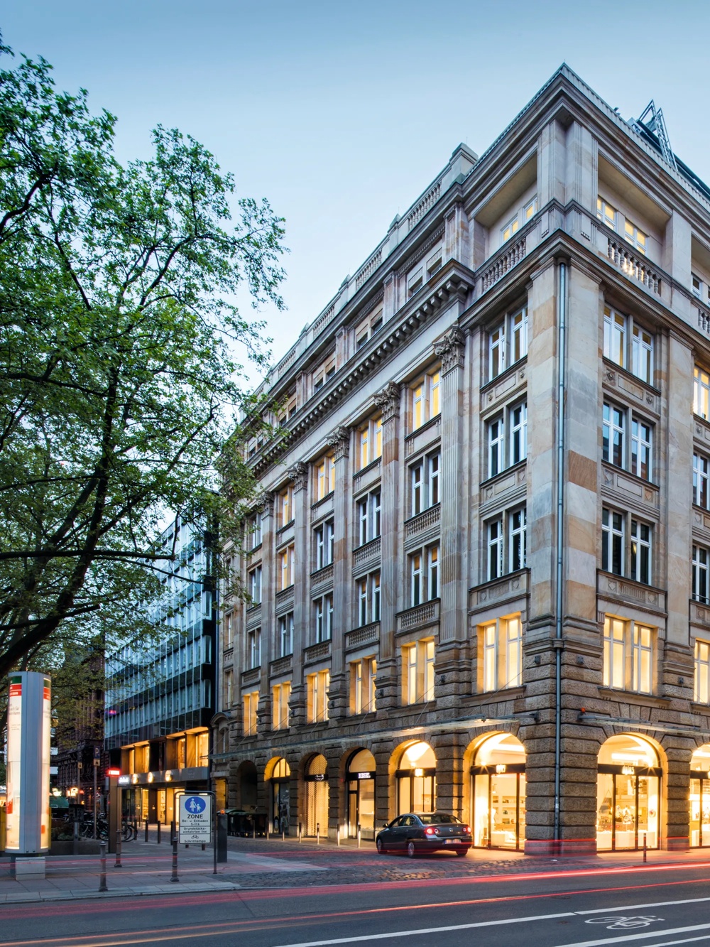 Eckgebäude mit beleuchteten Fenstern in der Dämmerung, Autos auf der Straße, Bäume links, moderner Anbau rechts.