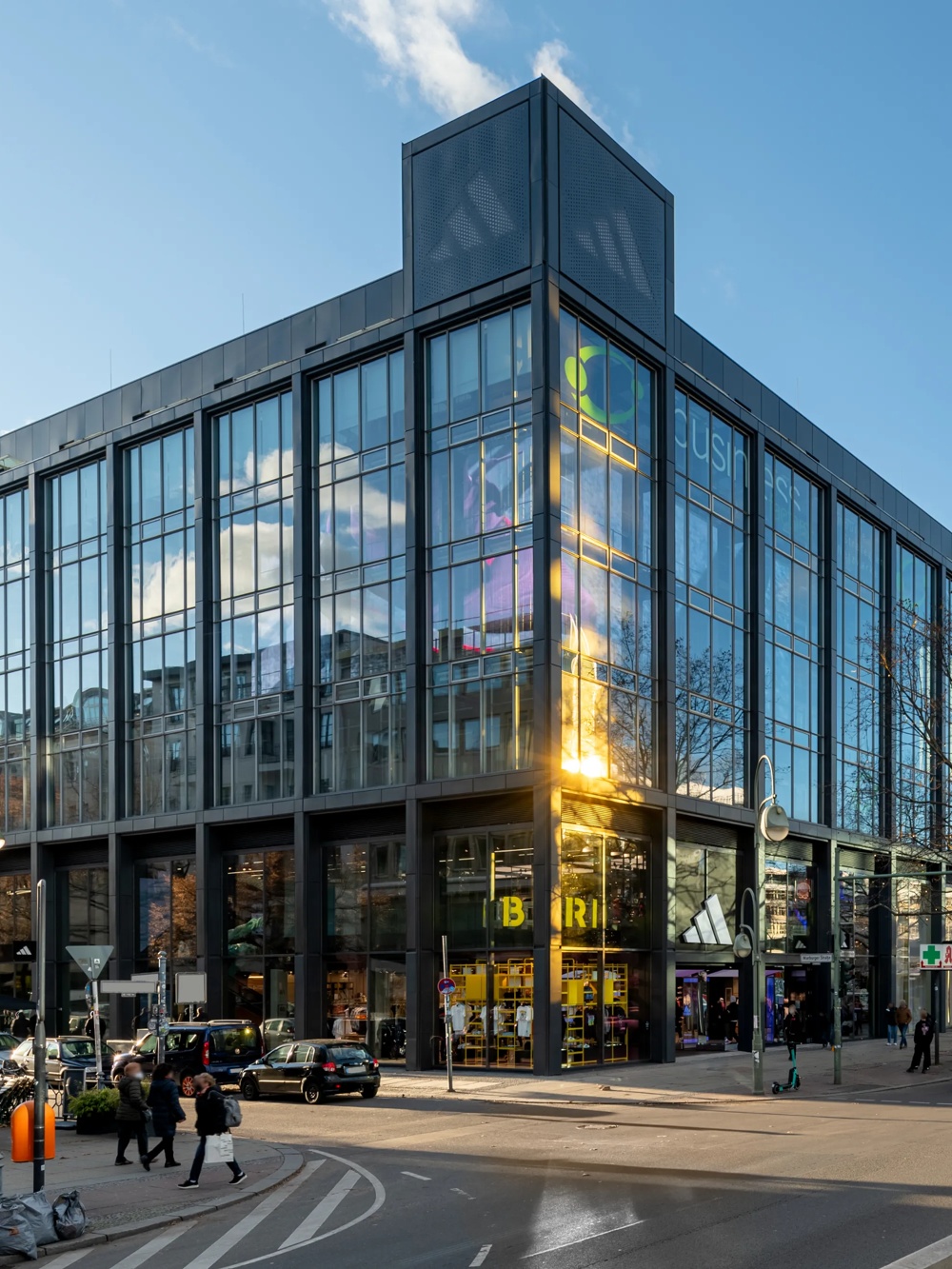 Moderne Glasfassade eines Gebäudes bei Sonnenschein, Menschen und Autos auf der Straße, blauer Himmel mit Wolken.