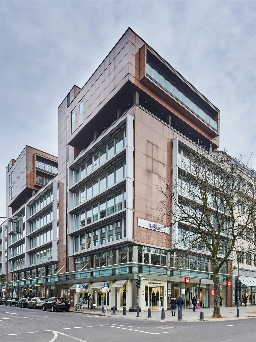 Straßenecke mit modernen Gebäuden, kahlen Bäumen und geparkten Autos unter bewölktem Himmel.