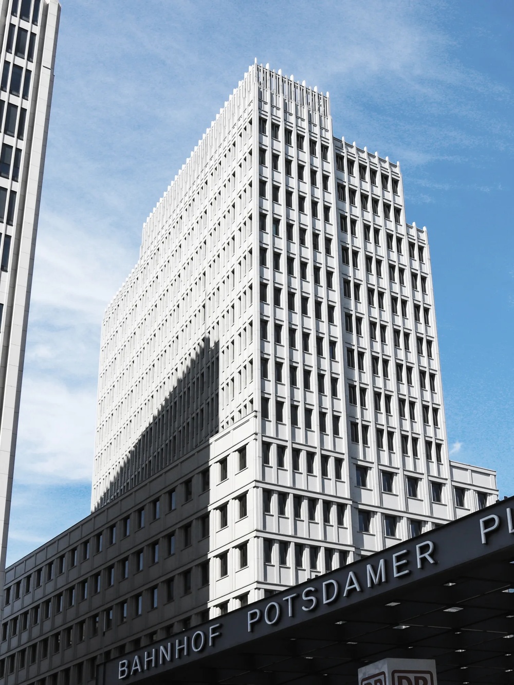 Ein modernes Hochhaus mit vielen Fenstern vor blauem Himmel, Schriftzug "Potsdamer Platz" im Vordergrund.