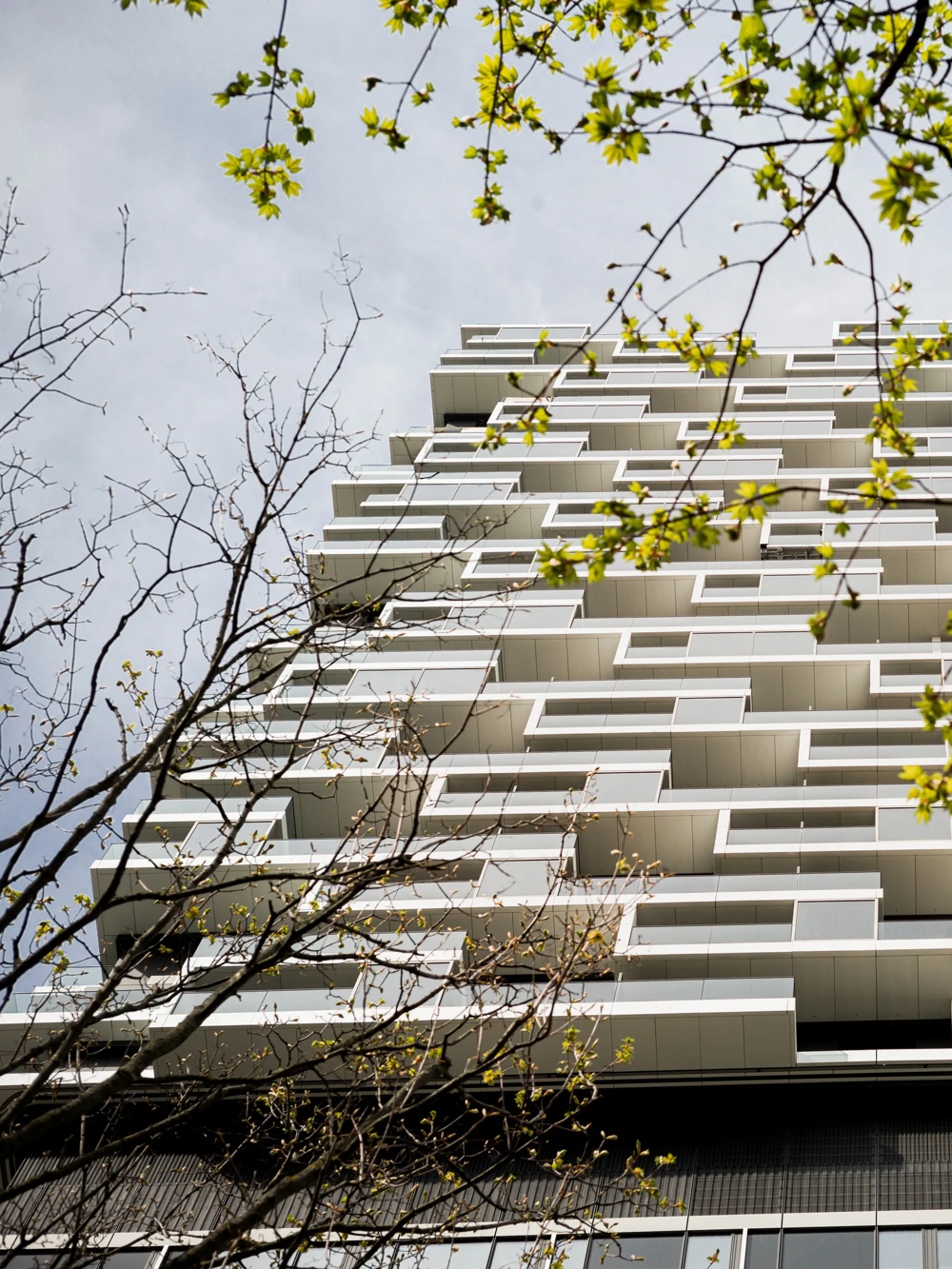 Ein modernes Hochhaus mit versetzten Balkonen, umgeben von blühenden Baumzweigen unter blauem Himmel.