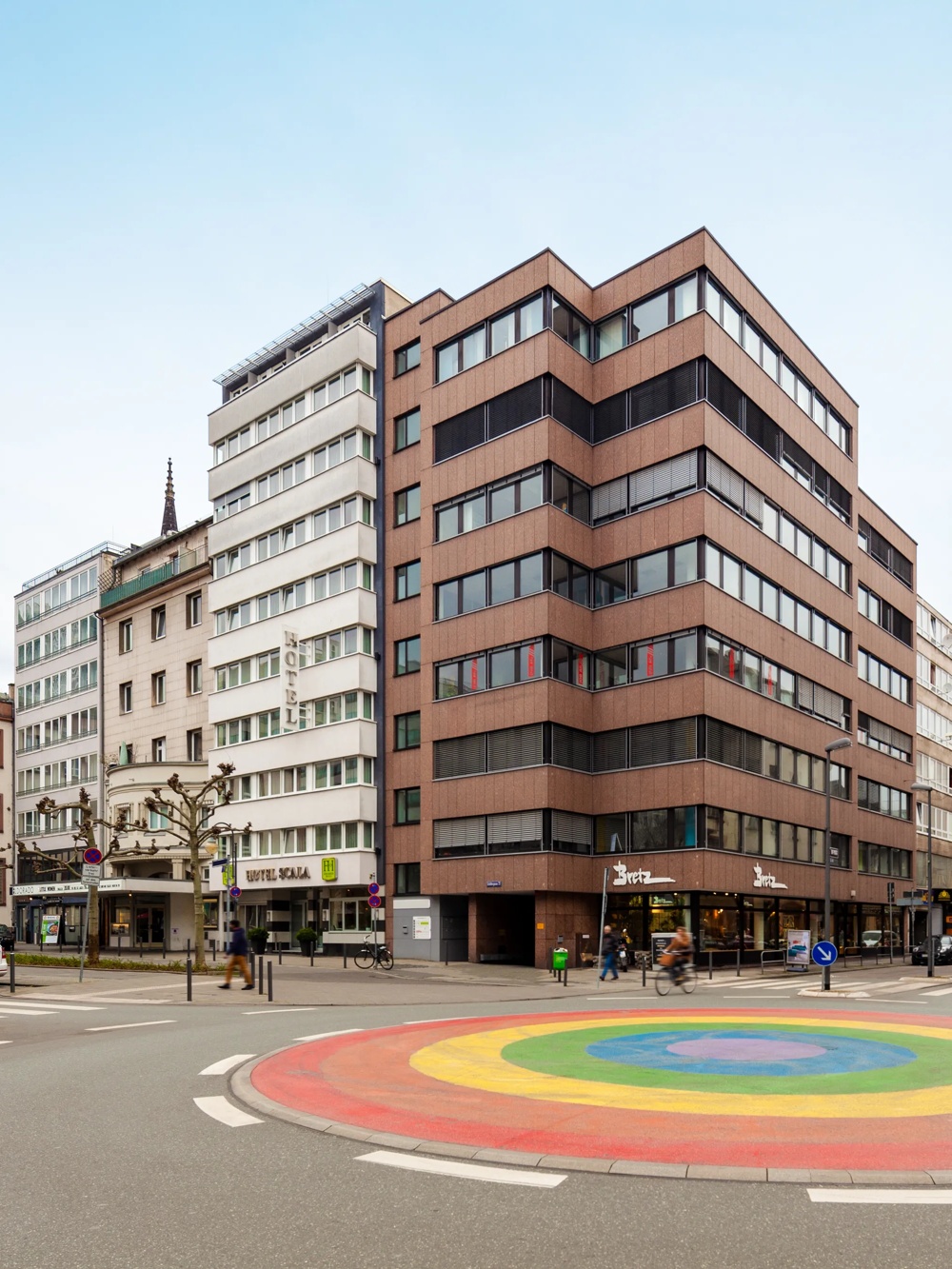 Ein Kreisverkehr mit Regenbogenfarben, umgeben von modernen Gebäuden und Geschäften in einer Stadtstraße.