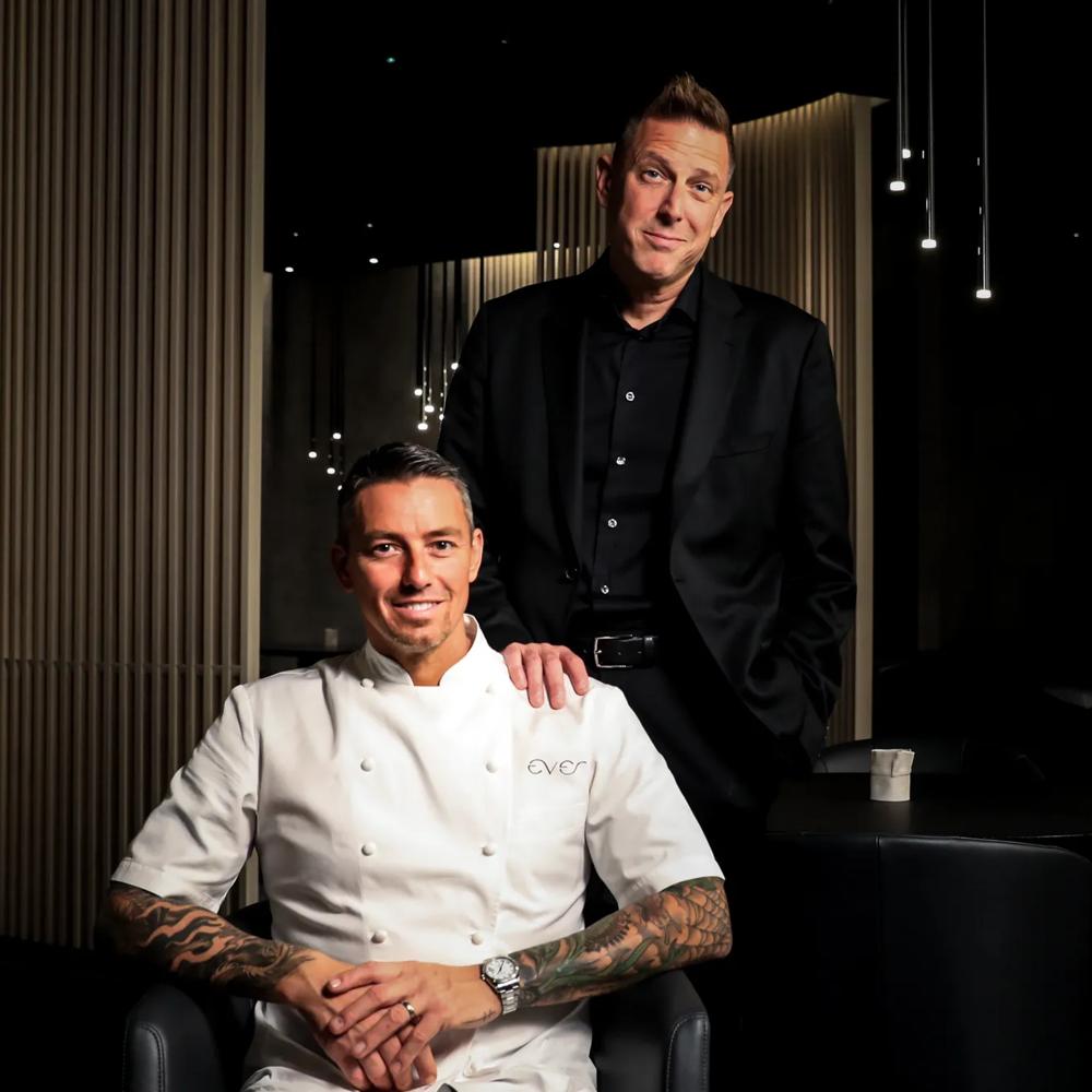 Ein tätowierter Koch in weißer Jacke sitzt, ein Mann in schwarzem Anzug steht daneben in einem eleganten, dunklen Raum.