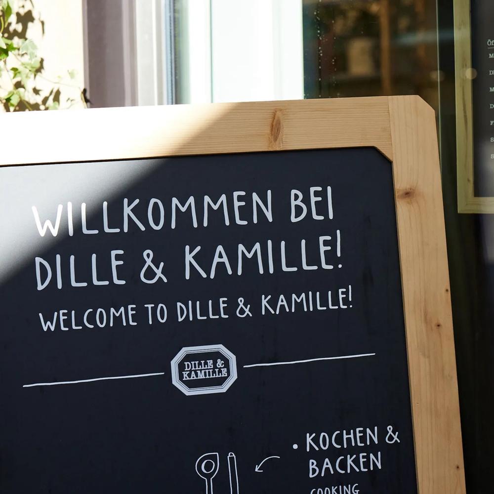 Willkommensschild von Dille & Kamille neben einem Fenster mit Öffnungszeiten und Pflanzen im Hintergrund.