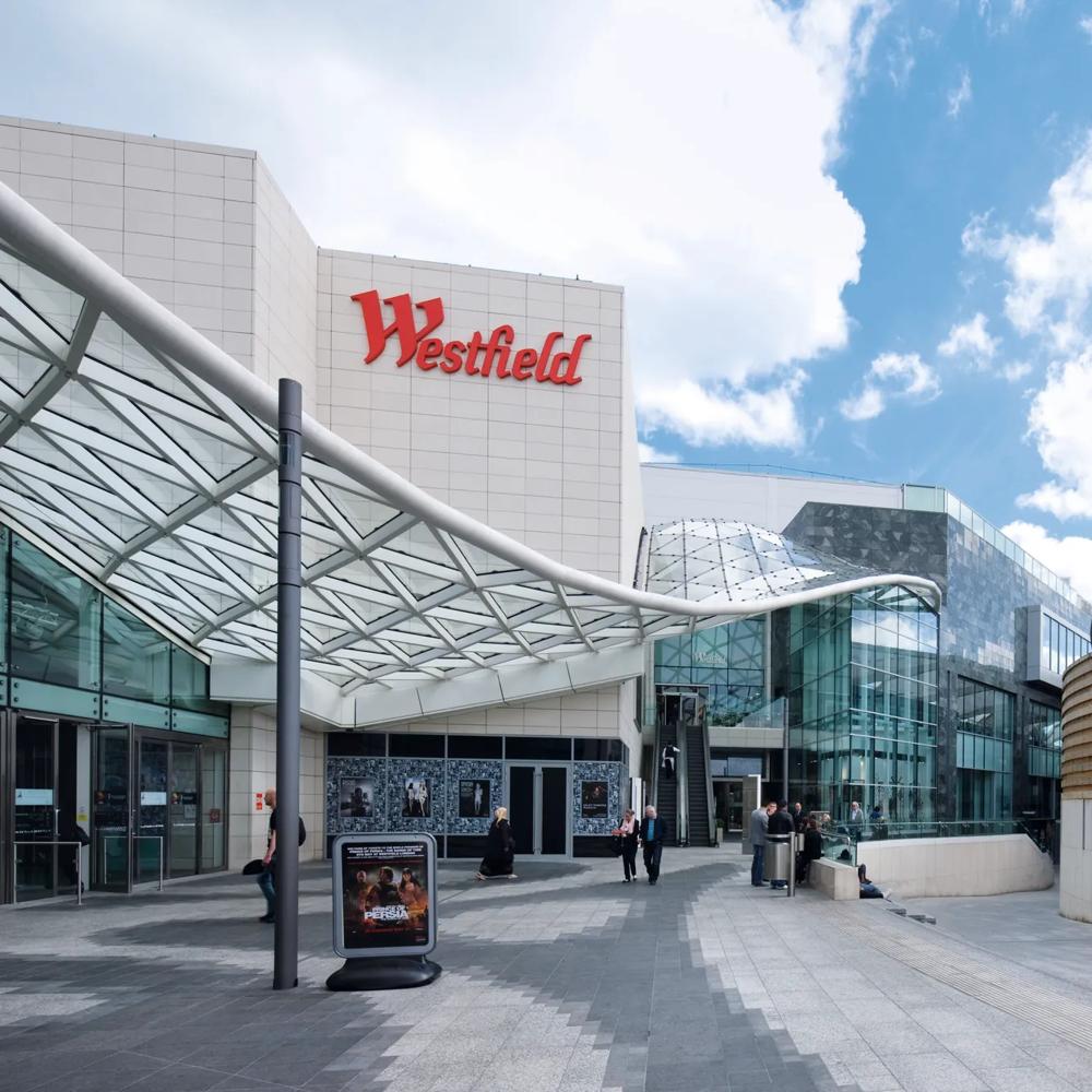 Eingang eines modernen Einkaufszentrums mit Glasdach und Westfield-Logo unter blauem Himmel.