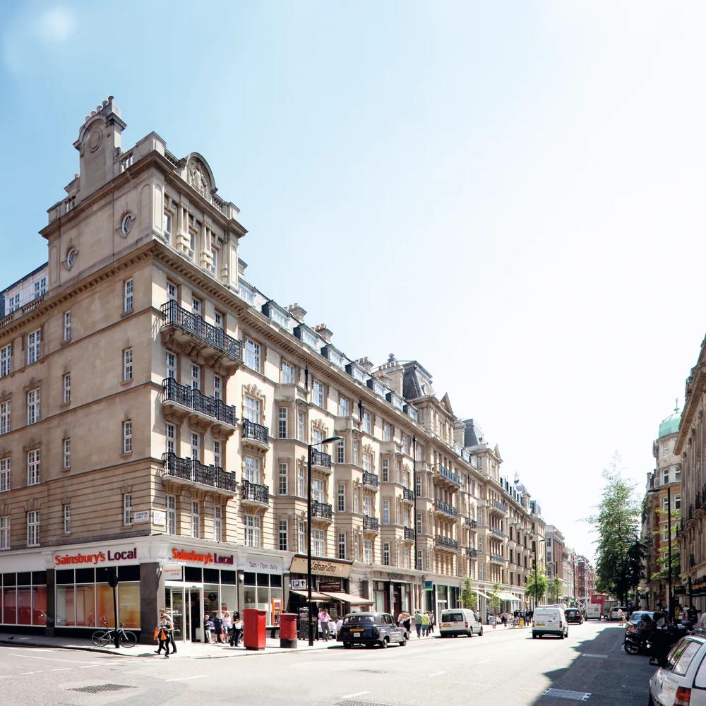 Straßenszene mit historischem Gebäude, Sainsbury's Local im Erdgeschoss, Menschen und Autos auf der Straße.