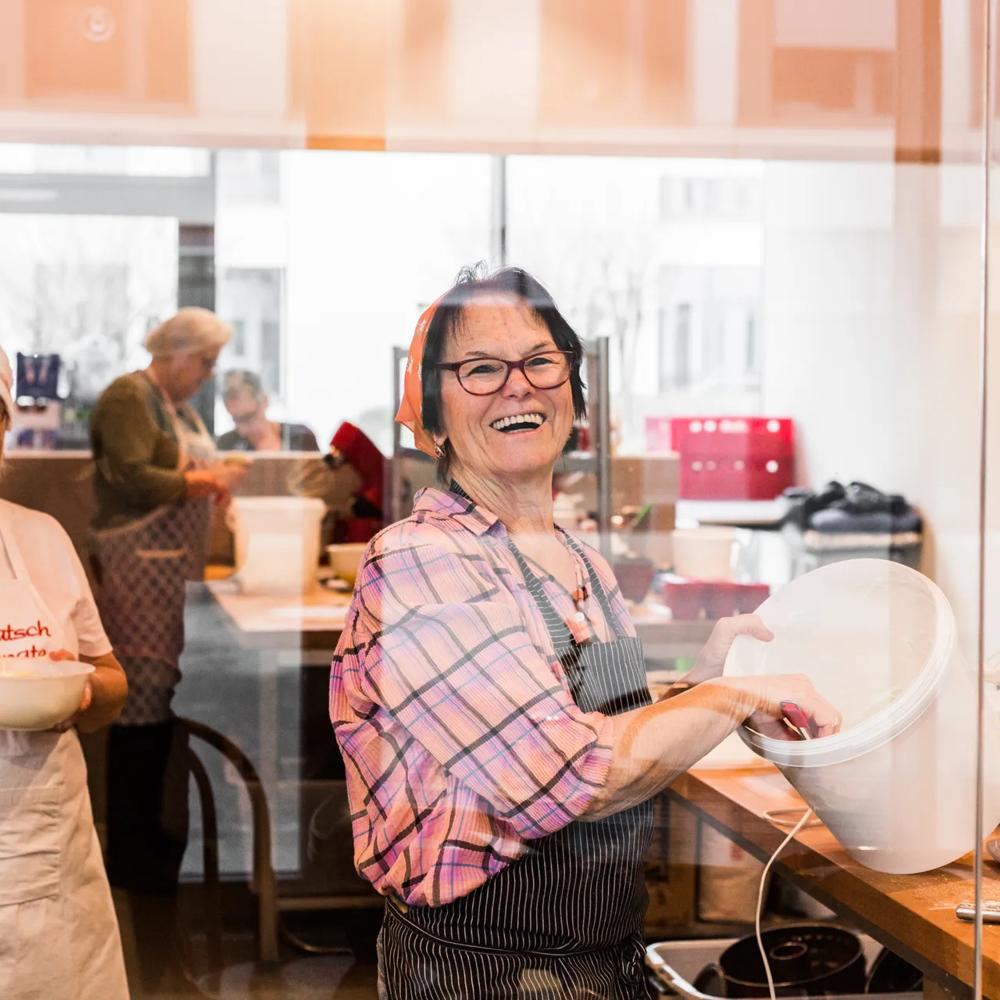 Zwei lachende Frauen in einer Bäckerei, eine winkt, die andere rührt Teig in einer Schüssel.