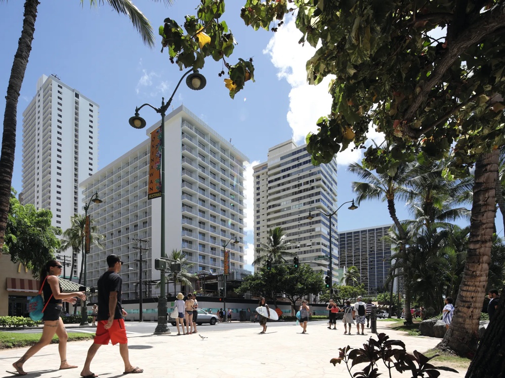 Straßenszene mit hohen Gebäuden, Palmen, Fußgängern und einem Surfer unter blauem Himmel.